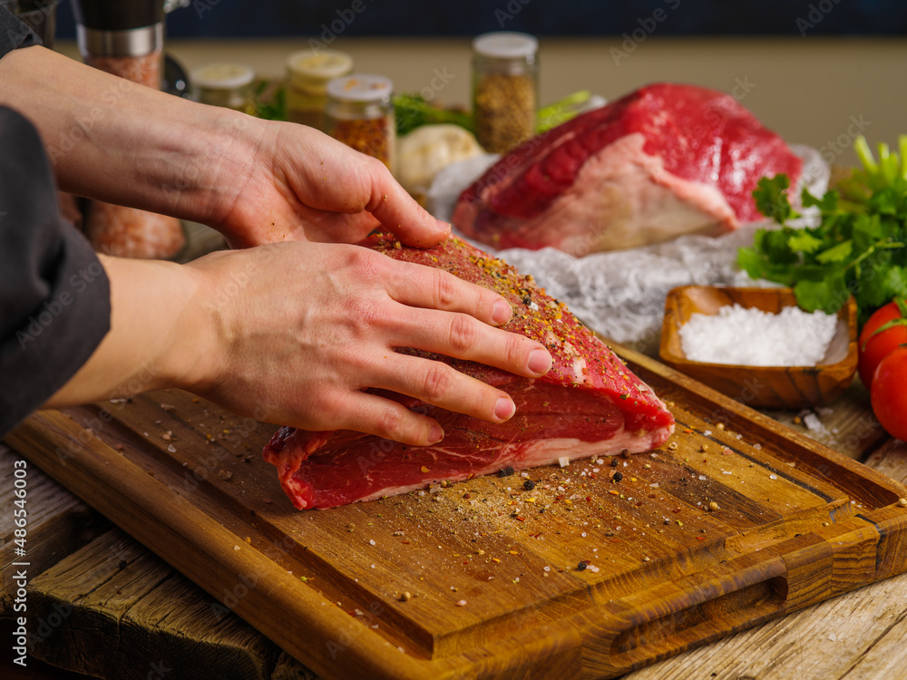 The chef's hands are seasoning a large piece of farm meat on a wooden cutting board. Cooking meat dishes in a restaurant and home kitchen, cookbook, culinary blog, advertising.