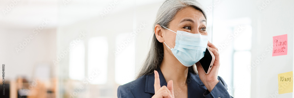 The happy Asian woman in protective mask talking on the phone and raising index finger up in the office The happy Asian woman in protective mask talking on the phone and raising index finger up in the office