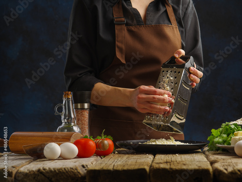 cooking dishes with cheese by the chef s hands in a professional kitchen on a wooden table and a blue background. The chef grates cheese. Recipes for dishes with cheese - pizza  salad  sauce.