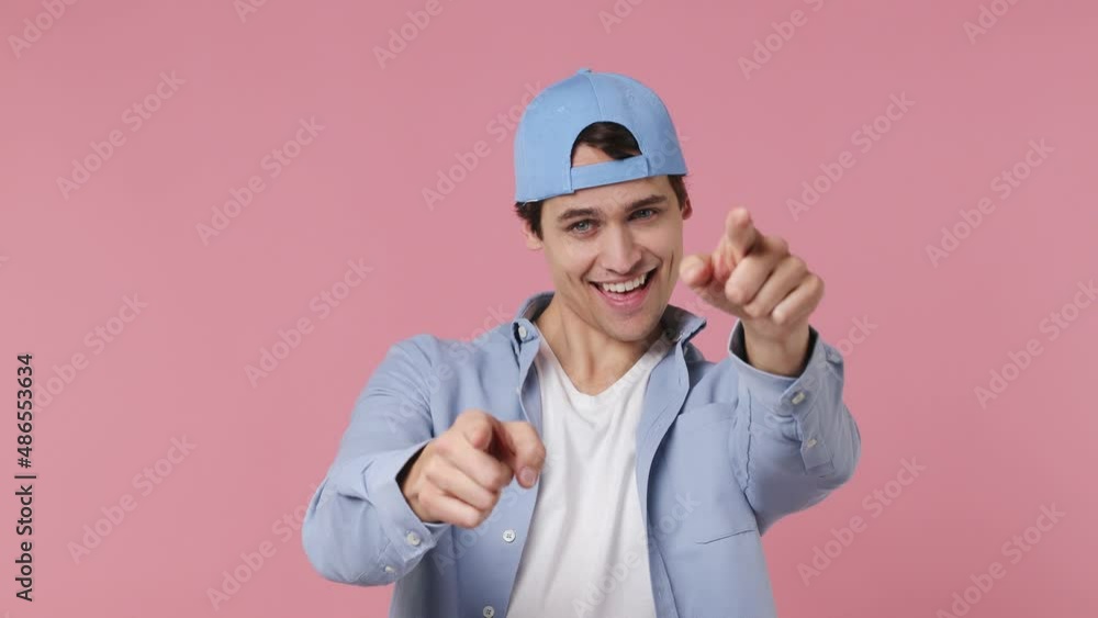 Happy blithesome charismatic fascinating fun young brunet man 20s years old wears blue shirt put on cap hat point index fingers camera on you dance isolated on plain pink background studio portrait Happy blithesome charismatic fascinating fun young brunet man 20s years old wears blue shirt put on cap hat point index fingers camera on you dance isolated on plain pink background studio portrait