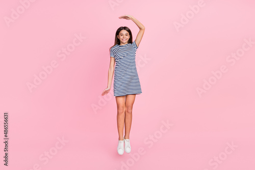Photo of cute charming girl wear striped dress jumping high measuring height isolated pink color background photo