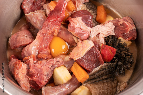 Closeup metal pet bowl with chicken necks, beef meat and bones, paunch, raw egg and carrot pieces. Natural organic daily meal ration preparation for dogs concept. Copy space.