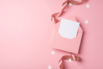 Top view photo of valentine's day decorations curly silk ribbon open pink envelope with paper card and heart shaped confetti on isolated pastel pink background with copyspace