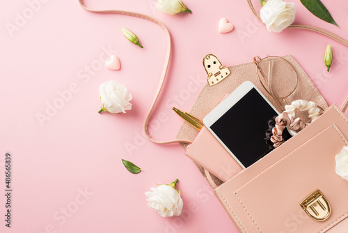 Top view photo of woman's day composition open pink leather bag with smartphone scrunchies glasses pen small hearts and prairie gentian flower buds on isolated pastel pink background with copyspace