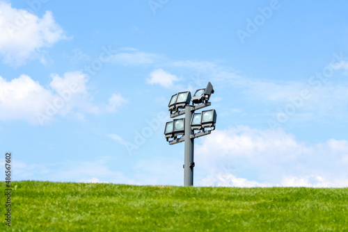 Electric spotlight on a metal pole during a sunny day on a green lawn. White clouds in the blue sky in the background
