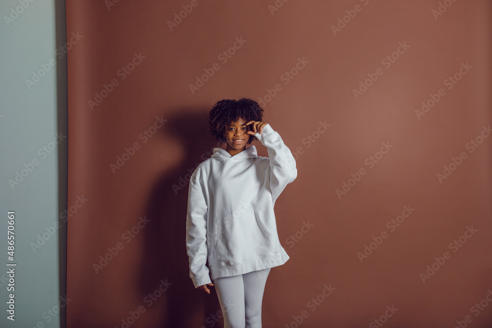 African american woman in a white hoodie and leggings stands on a brown background. Mock-up. African american woman in a white hoodie and leggings stands on a brown background. Mock-up.