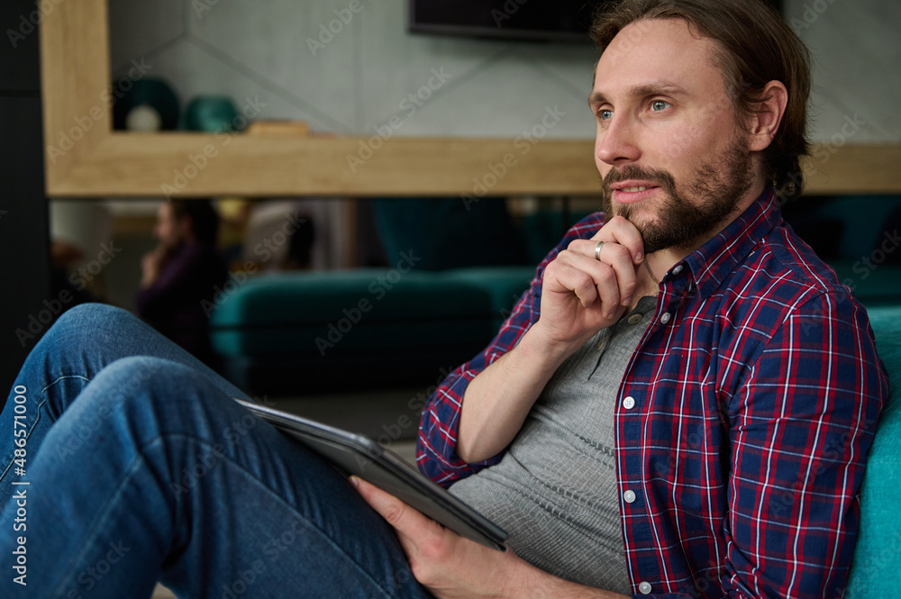 Pensive handsome Caucasian man in casual denim and checkered shirt holding a digital tablet and looking thoughtfully away, sitting at home environment Pensive handsome Caucasian man in casual denim and checkered shirt holding a digital tablet and looking thoughtfully away, sitting at home environment