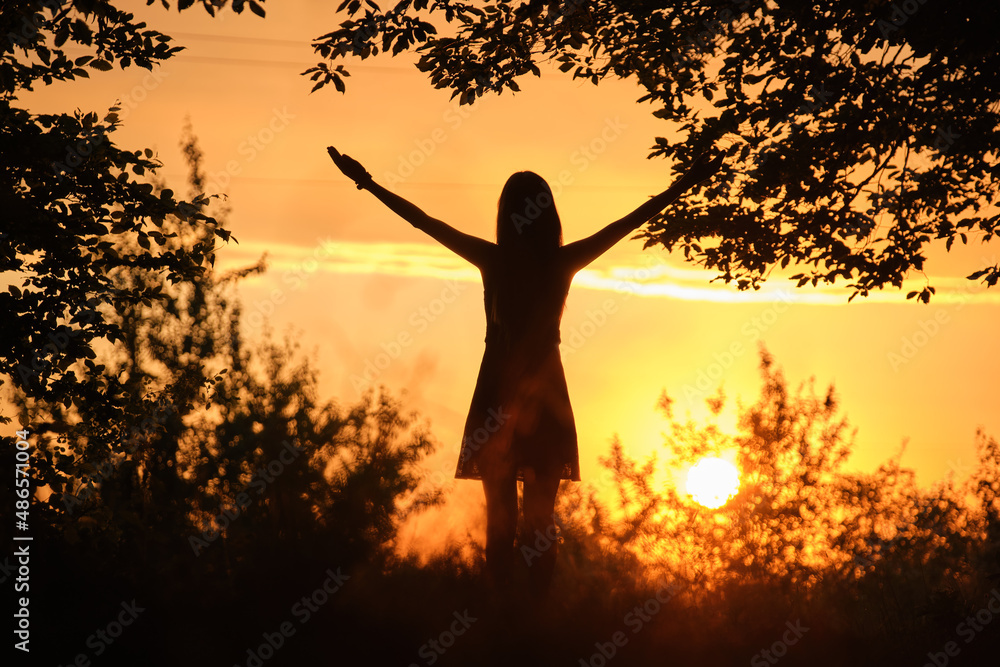 Rear silhouette view of young happy woman standing alone in dark woods with raised up hands enjoying summer evening. Victory and success concept