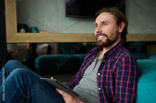 Relaxed smiling European guy in casual denim and checkered shirt reads news on digital tablet, surfs internet, sitting on floor at home leaning back on a sofa. Leisure time, recreation, lifestyle