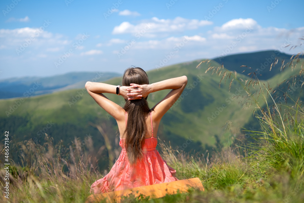 Young happy woman traveler in red dress resting on green grassy hillside on a windy day in summer mountains enjoying view of nature
