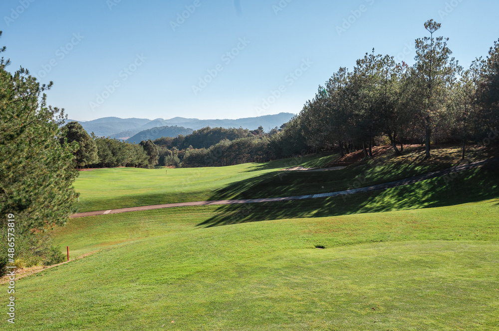 Golf course landscape with short green grass, some hills and trees.
