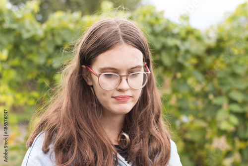 A beautiful girl with braces on her teeth clenching them looks down. Hipster teenager in glasses with long hair against blurred green foliage