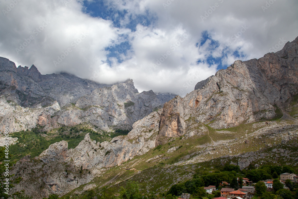 Fototapeta premium View of the rocky peaks of the Pyrenees, covered with low clouds. Green trees and roofs of houses in the foreground.