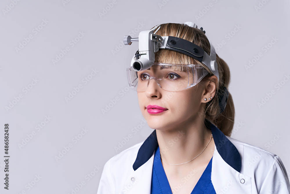 Confident ENT doctor wearing surgical headlight head light and protective glasses. Closeup portrait of female otolaryngologist or head and neck surgeon on light grey background.