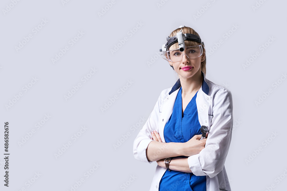 Confident ENT doctor wearing surgical headlight head light and protective glasses. Portrait of female otolaryngologist or head and neck surgeon standing arms crossed on light grey background.