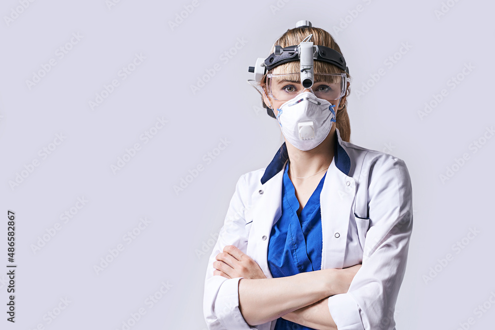 Confident ENT doctor wearing surgical headlight head lantern, protective face mask and glasses. Portrait of female otolaryngologist standing arms crossed on light grey background.