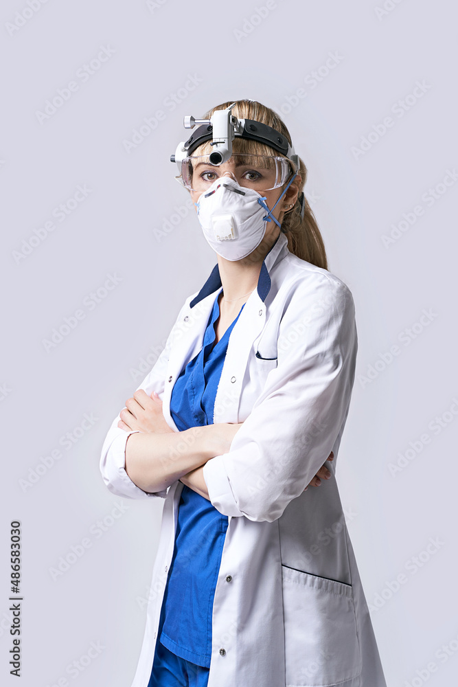 Confident ENT doctor wearing surgical headlight head lantern, protective face mask and glasses. Portrait of female otolaryngologist standing arms crossed on light grey background.