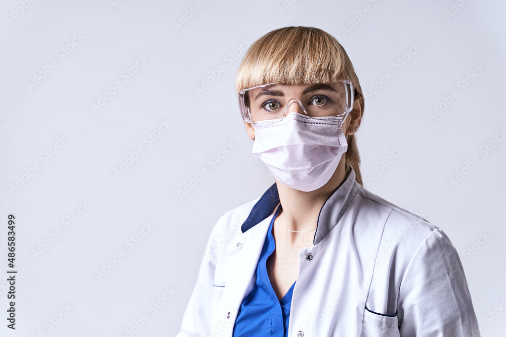 Confident blonde female doctor wearing protective face mask. Portrait on light grey background.