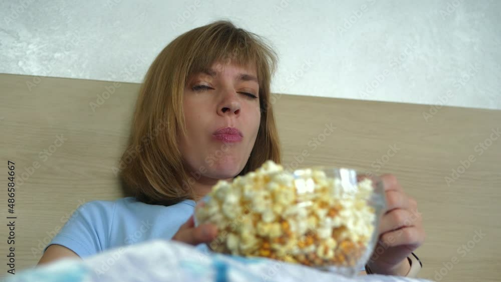 Girl actively eats popcorn from glass vase close-up. Woman watches TV, changes channels, lying in bed with popcorn. Relaxing at home alone Girl actively eats popcorn from glass vase close-up. Woman watches TV, changes channels, lying in bed with popcorn. Relaxing at home alone