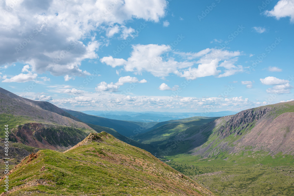 Scenic view from mountain ridge to green forest valley among mountain ranges and hills on horizon at changeable weather. Green landscape with sunlit mountain vastness under cumulus clouds in blue sky.