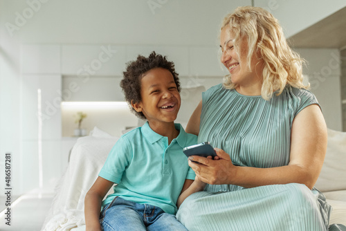 Happy mother or nanny resting on sofa using modern cellphone gadget with small boy child
