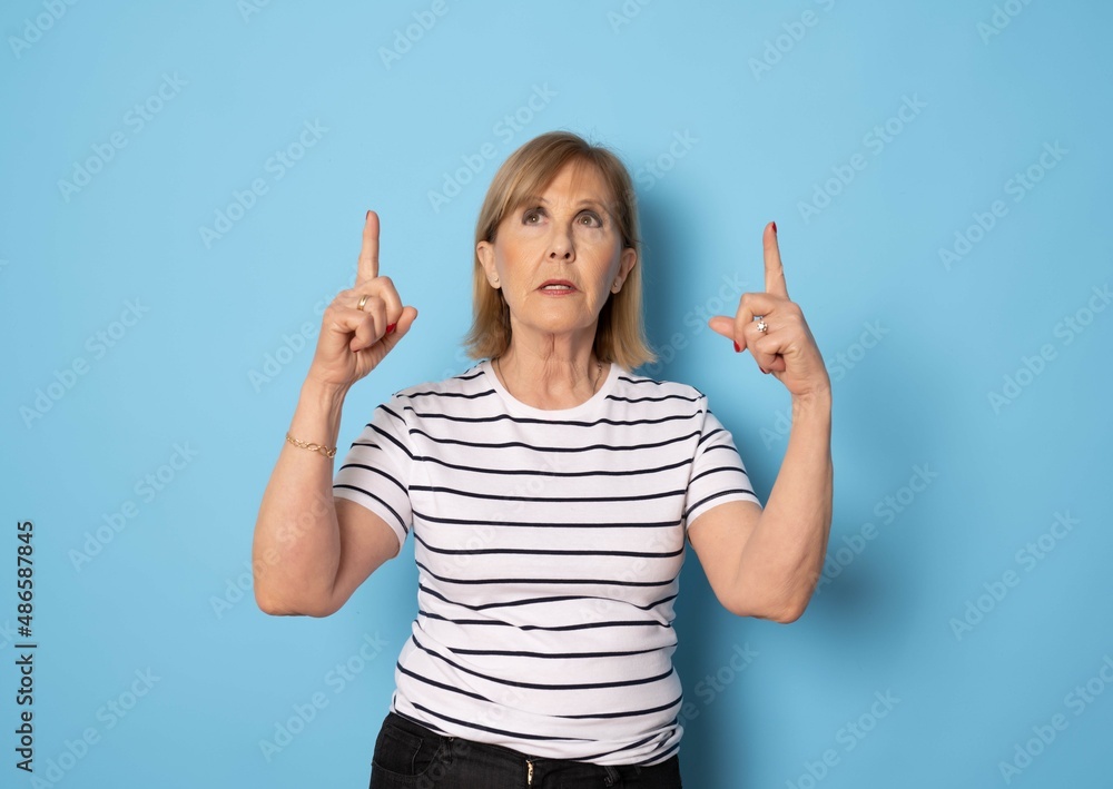 Senior caucasian woman in striped t-shirt pointing fingers up standing isolated over blue background. Senior caucasian woman in striped t-shirt pointing fingers up standing isolated over blue background.