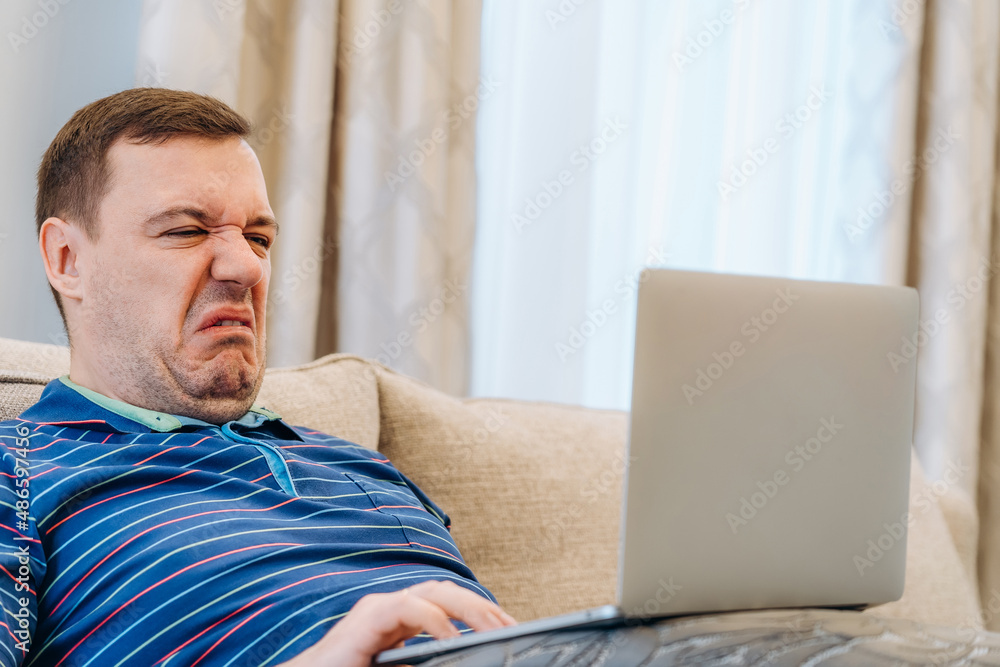 Fototapeta premium Annoyed millennial man with disgust confused looking at laptop screen sitting on sofa at living room. cringe guy looking at computer with squeamishness, reacting on watching disgusting video at home