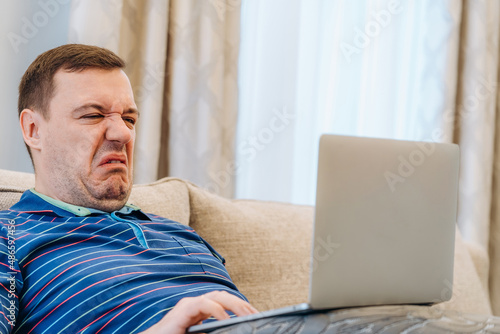 Annoyed millennial man with disgust confused looking at laptop screen sitting on sofa at living room. cringe guy looking at computer with squeamishness, reacting on watching disgusting video at home photo