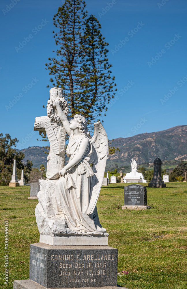Santa Barbara, California, USA - February 8, 2022: Calvary Cemetery. Closeup of white stone Edmund Arellanes unique historic tombstone on green burial lawn under blue sky. Tall tree