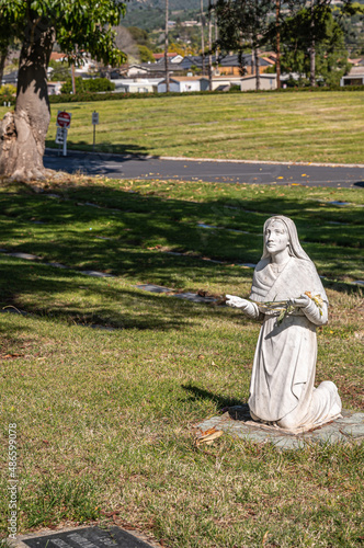 Santa Barbara, California, USA - February 8, 2022: Calvary Cemetery. Closeup of  Bernadette of Lourdes statue on green burial lawn. Faded housing buildings in back. photo