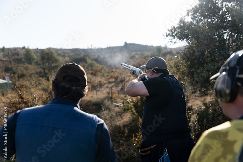 Canvastavla An adult man is shooting during a skeet shooting competition while two more part