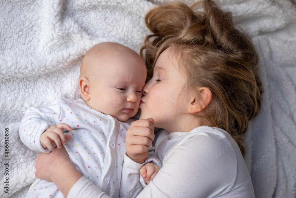 Happy children, toddler and older sister, hugging at home on a white blanket, smiling, shot from above. blonde girl hugging her newborn sister Happy children, toddler and older sister, hugging at home on a white blanket, smiling, shot from above. blonde girl hugging her newborn sister