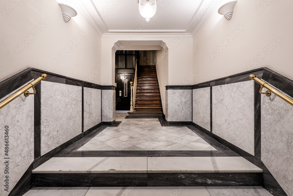 Fototapeta premium Entrance hall of a vintage apartment residential building with wooden staircase, white veined marble tiled half wall and elevator