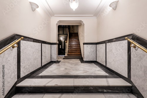 Entrance hall of a vintage apartment residential building with wooden staircase, white veined marble tiled half wall and elevator