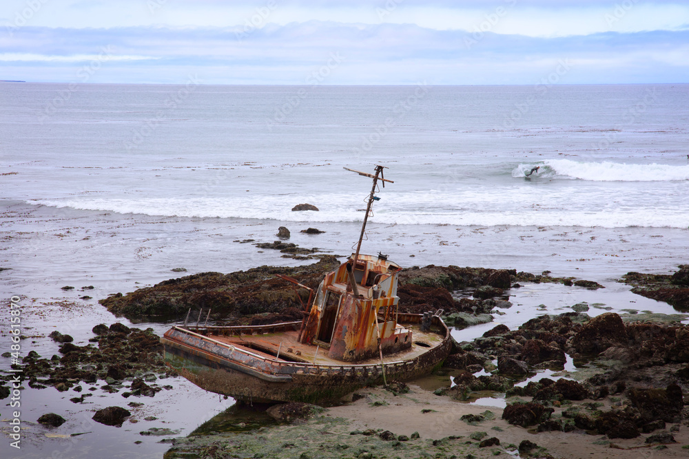 Old, rusty fishing vessel shipwrecked, beached on the rocks while surfer catches a wave on a gray day at the beach in Cayucos, California