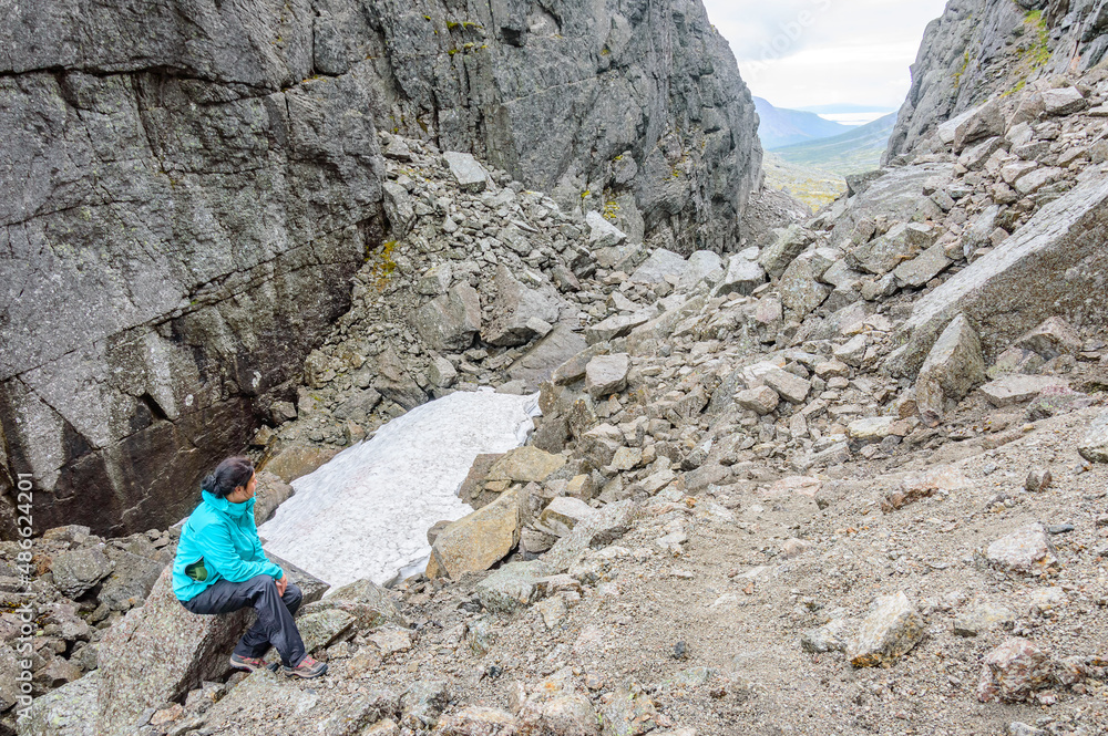 Young hiker woman in turquoise jacket and black trousers sitting on a stone in the mountains. White snow, grey rocks and stones