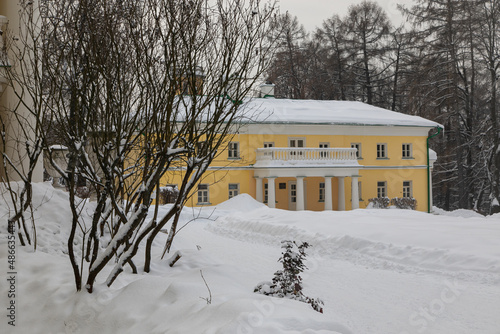 A vintage yellow manor house with white columns stands in a winter garden. It is surrounded by trees and a lot of snow. The drifts are deep and the trees are covered with snow. © Vadim