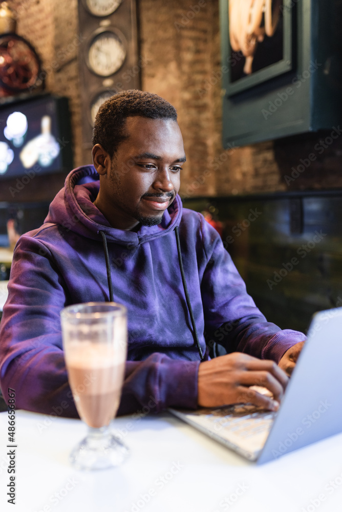 Young African American male freelancer in stylish hoodie sitting at table and typing on netbook and having glass of chocolate milkshake in cafe