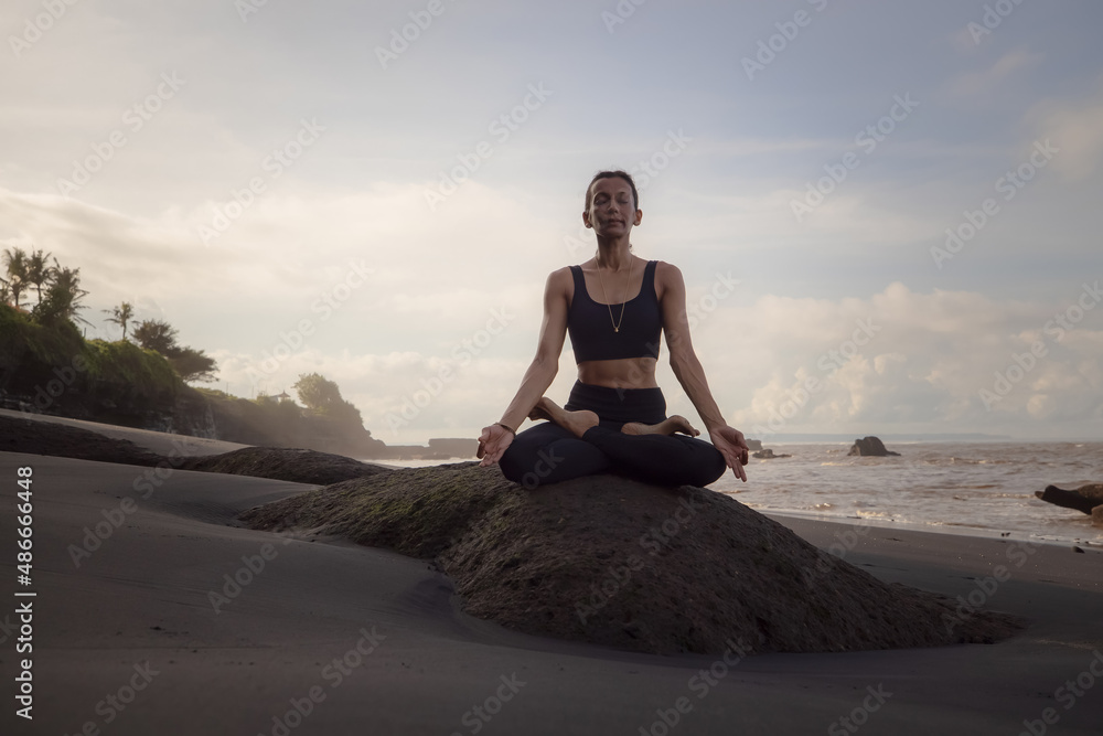 Meditation yoga on the beach. Asian woman sitting on the rock in Lotus pose. Padmasana. Hands in gyan mudra. Yoga retreat. Closed eyes. Healthy concept. Copy space. Mengening beach, Bali