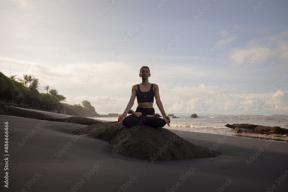 Fototapeta premium Morning meditation and yoga on the beach. Asian woman sitting on the rock in Lotus pose. Padmasana. Hands in gyan mudra. Yoga retreat. Healthy concept. Copy space. Mengening beach, Bali