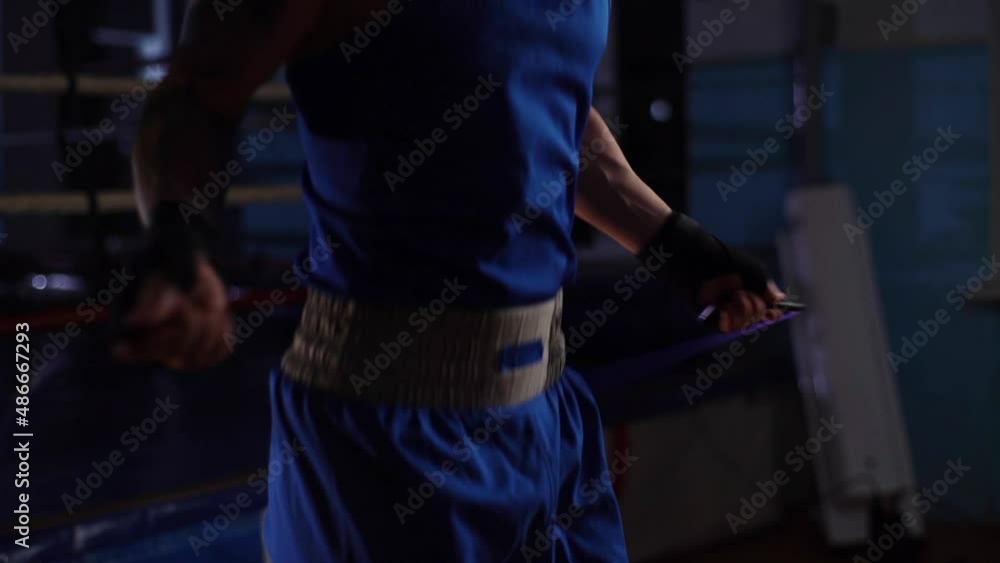 Close-up cropped shot of torso unrecognizable male boxer with strong body jumping with skipping rope during workout training, warming up before boxing workout in sport club with dark interior.