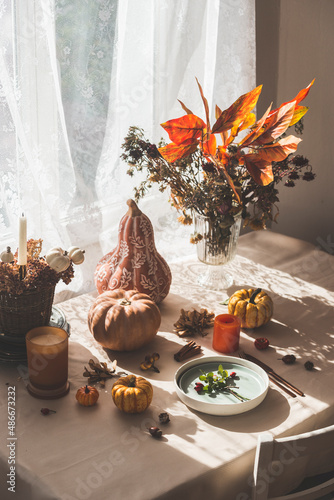 Autumn still life with pumpkins, leaves bunch and candle at table and window background with white curtains and sunlight. Cozy indoor scene in autumn. Front view.
