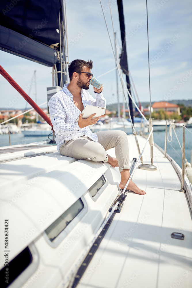 A young man with a tablet is enjoying a coffee while sitting on a yacht and riding through the dock on the seaside. Summer, sea, vacation A young man with a tablet is enjoying a coffee while sitting on a yacht and riding through the dock on the seaside. Summer, sea, vacation
