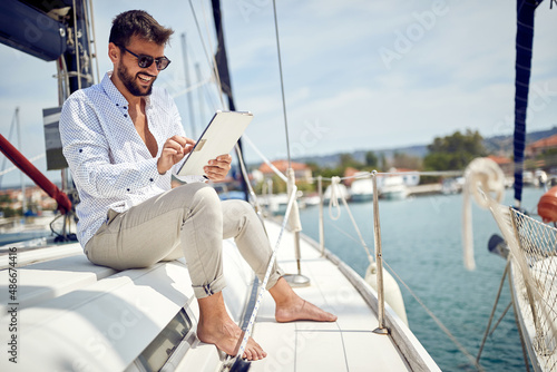 A young man enjoying a tablet content while sitting on a yacht and riding in the dock on the seaside. Summer, sea, vacation © luckybusiness