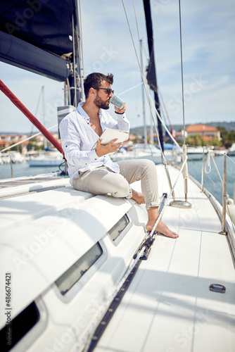 A young man with a tablet is enjoying a coffee while sitting on a yacht and riding through the dock on the seaside. Summer, sea, vacation