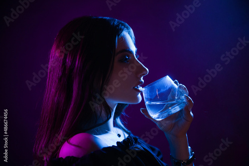 A young woman in the dark in a club drinks from a glass. Party girl in blue and pink light. Beautiful model portrait in cyber style with color music