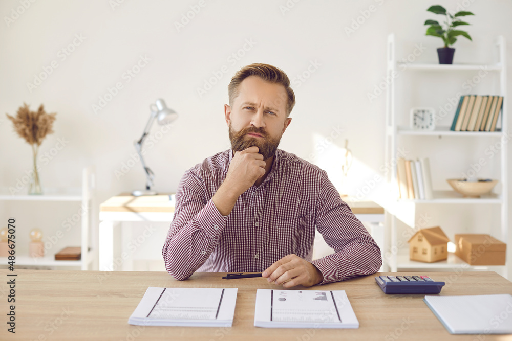 Portrait of focused pensive businessman looking at camera while sitting in office. Man sits at table with documents and calculator and thinks about plan to increase company's profits. Portrait of focused pensive businessman looking at camera while sitting in office. Man sits at table with documents and calculator and thinks about plan to increase company's profits.