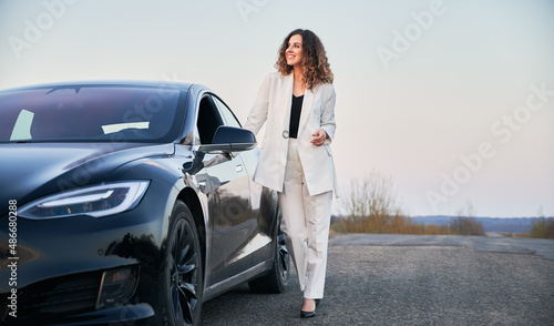 Young businesswoman in stylish white suit posing near modern electric car on roadside. © anatoliy_gleb