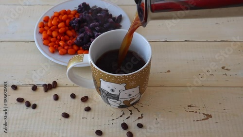 From the coffee maker, black coffee is poured into a cup, coffee beans are scattered nearby and bright berries are in a white plate. Slow motion video.