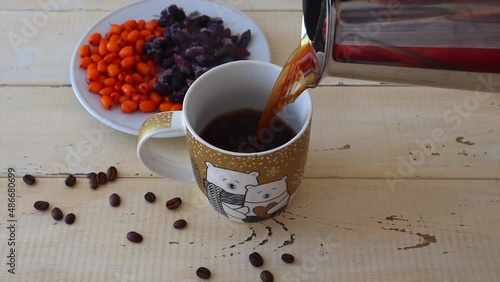 From the coffee maker, black coffee is poured into a cup, coffee beans are scattered nearby and bright berries are in a white plate.